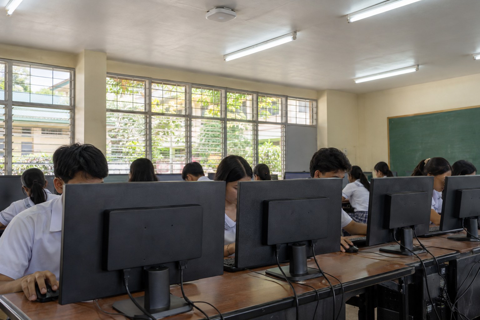 Students in white school uniforms work at rows of desktop computers in a sunlit public school computer lab, with a Wi-Fi access point mounted on the ceiling.